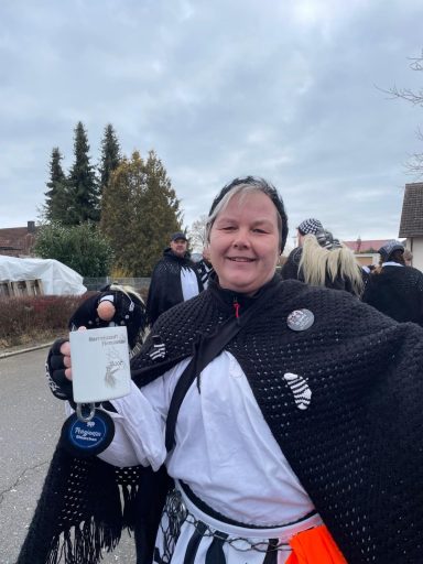 Frau mit Umhang hält ein Schild und lächelt, umgeben von anderen Personen.