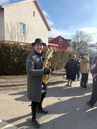 Mann mit einem Saxophon, gekleidet in einem grauen Mantel und Hut, in einer Menschenmenge.