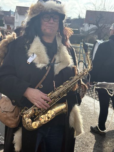 Person mit Saxophon in einem Pelzmantel, umgeben von anderen Musikern im Freien.