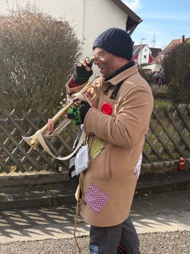 Mann in beiger Jacke mit Trompete, telefoniert und trägt bunte Patches.