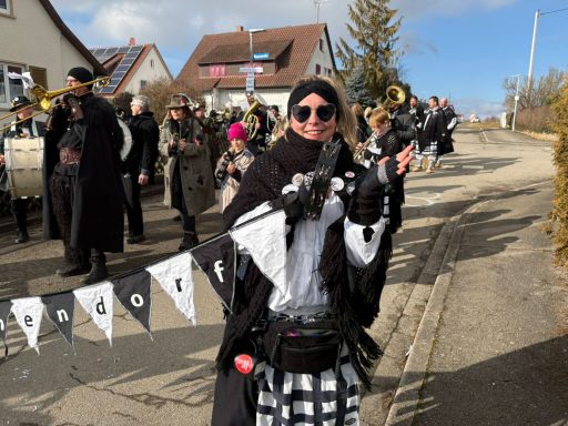 Eine Person in schwarzer Kleidung posiert vor einer Gruppe von festlich gekleideten Menschen.