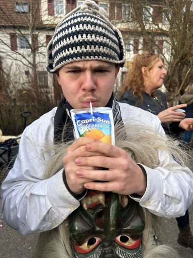 Junger Mann mit gestreiftem Hut, trinkt Saft, hält eine Maske in der anderen Hand.