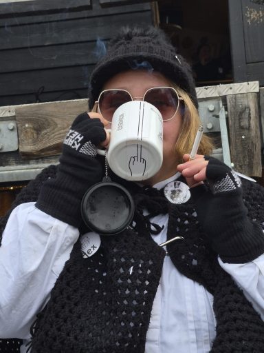 Person in warmer clothing mit Sonnenbrille, hält Tasse und Zigarette.