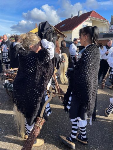 Personen in traditioneller Kleidung mit schwarz-weißen Streifen und Umhängen, nahe einem Pferd.