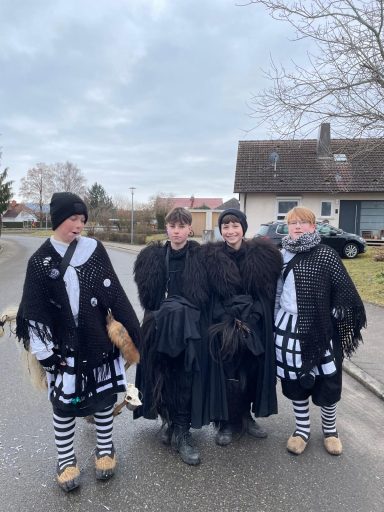 Vier Personen in auffälligen Kostümen mit schwarzen Umhängen und gestreiften Strümpfen.