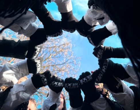 Hände in schwarzen Handschuhen bilden einen Kreis unter blauem Himmel.