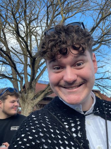 Junger Mann mit lockigem Haar und einem breiten Grinsen steht vor einem Baum.