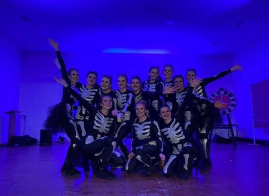 Tanzgruppe in schwarzen Kostümen mit Skeletdesign vor blauem Hintergrund.