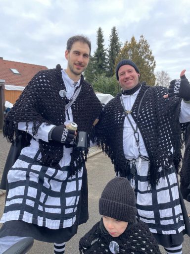 Zwei Männer in schwarz-weißen Kostümen mit Schals und Mützen, lächeln und posieren.