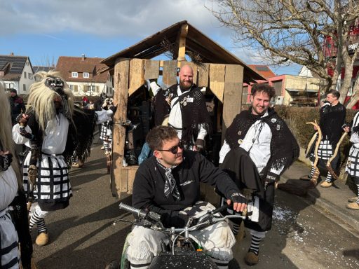 Männer in schwarz-weißen Kostümen feiern Karneval vor einem Holzaufbau.