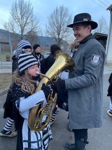Ein Junge mit einer Tuba steht neben einem Mann in einem grauen Mantel. Beide tragen Streifen.