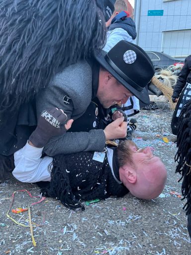 Zwei Männer in Kostümen kämpfen spielerisch am Boden. Konfetti verstreut.