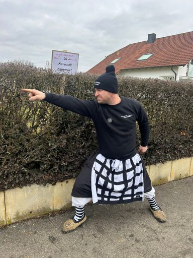 Mann in sportlicher Kleidung und Hut mit ausgefahrenem Arm steht vor einer Hecke.